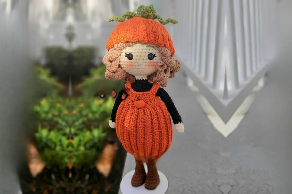 Uma boneca de crochê com roupa laranja e segurando abóboras