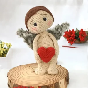Anjinho em amigurumi bege com cabelos castanhos, asas cinza e coração vermelho