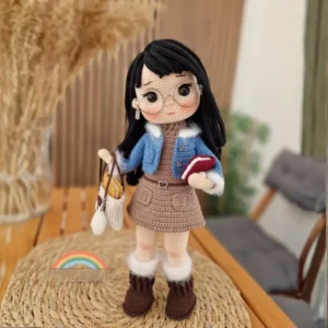 Boneca Clara em amigurumi com cabelos pretos, óculos redondos, jaqueta azul, saia marrom e botas