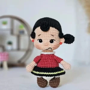 Boneca menina em amigurumi com cabelos pretos, roupa vermelha, saia preta com detalhe amarelo e sapatos marrons