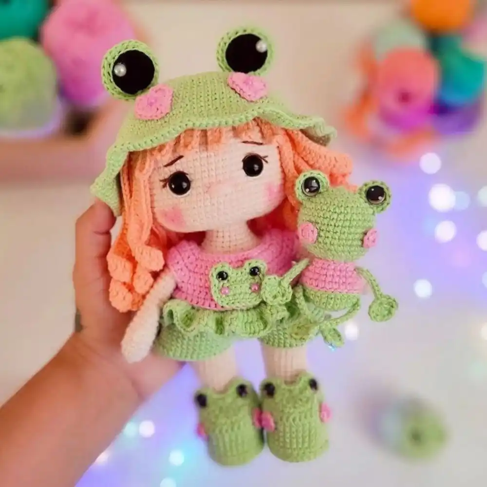 Boneca em amigurumi com cabelos laranja, chapéu verde de sapo e pequeno sapinho nos braços