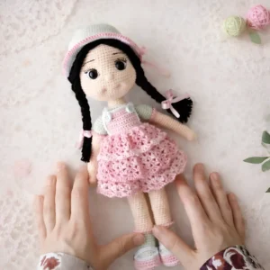 Boneca em amigurumi com cabelos pretos em duas tranças, gorrinho claro e vestido rosa
