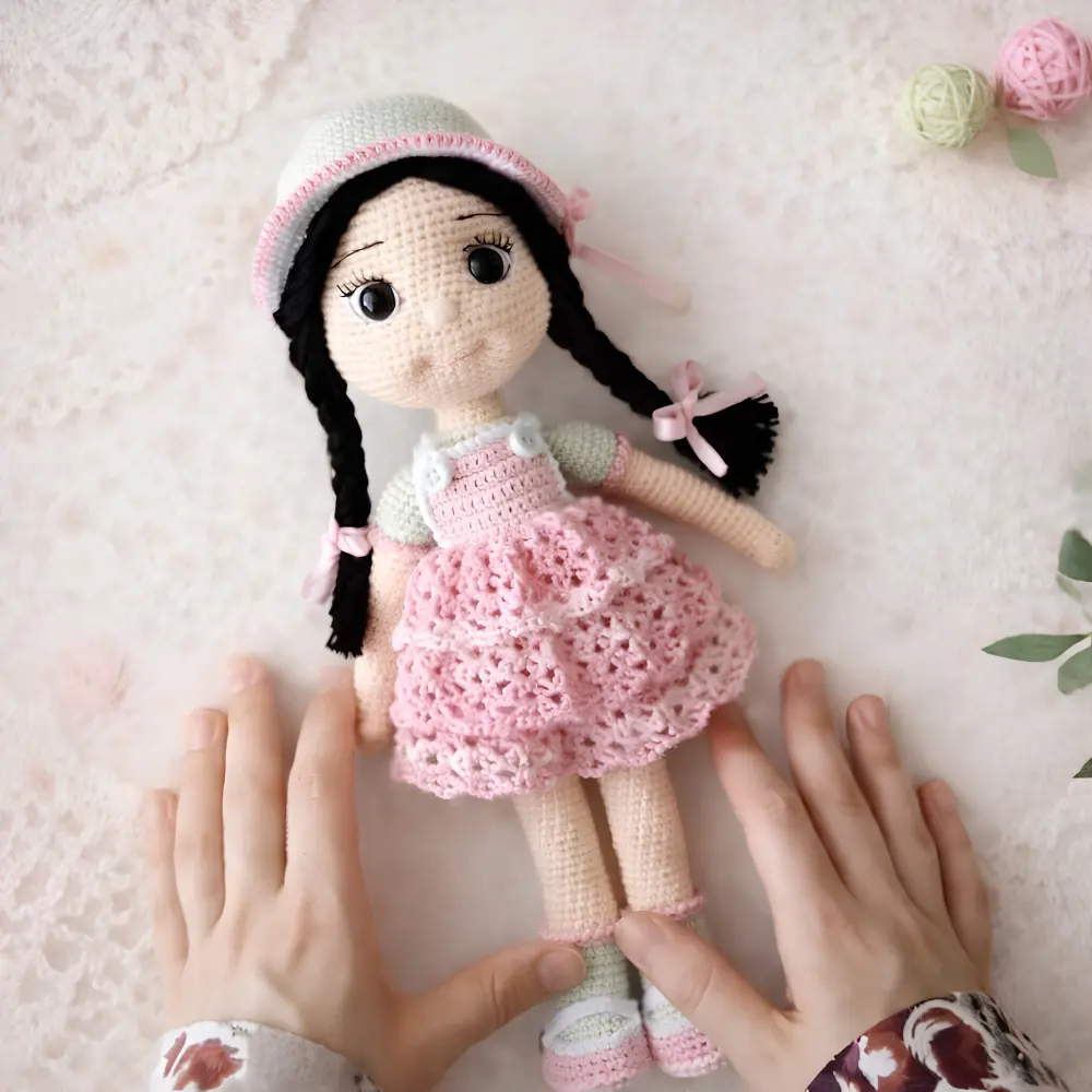Boneca em amigurumi com cabelos pretos em duas tranças, gorrinho claro e vestido rosa