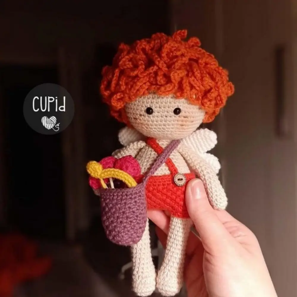 Boneco Cupido em amigurumi com cabelo ruivo cacheado, asas brancas, roupa vermelha e bolsa de corações