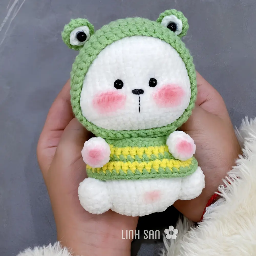 Bonequinho branco em amigurumi com capuz verde de sapo e roupa listrada verde e amarela