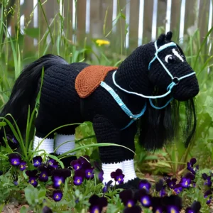 Cavalo preto em amigurumi com sela marrom, acessórios azul-claro e patas com detalhes brancos