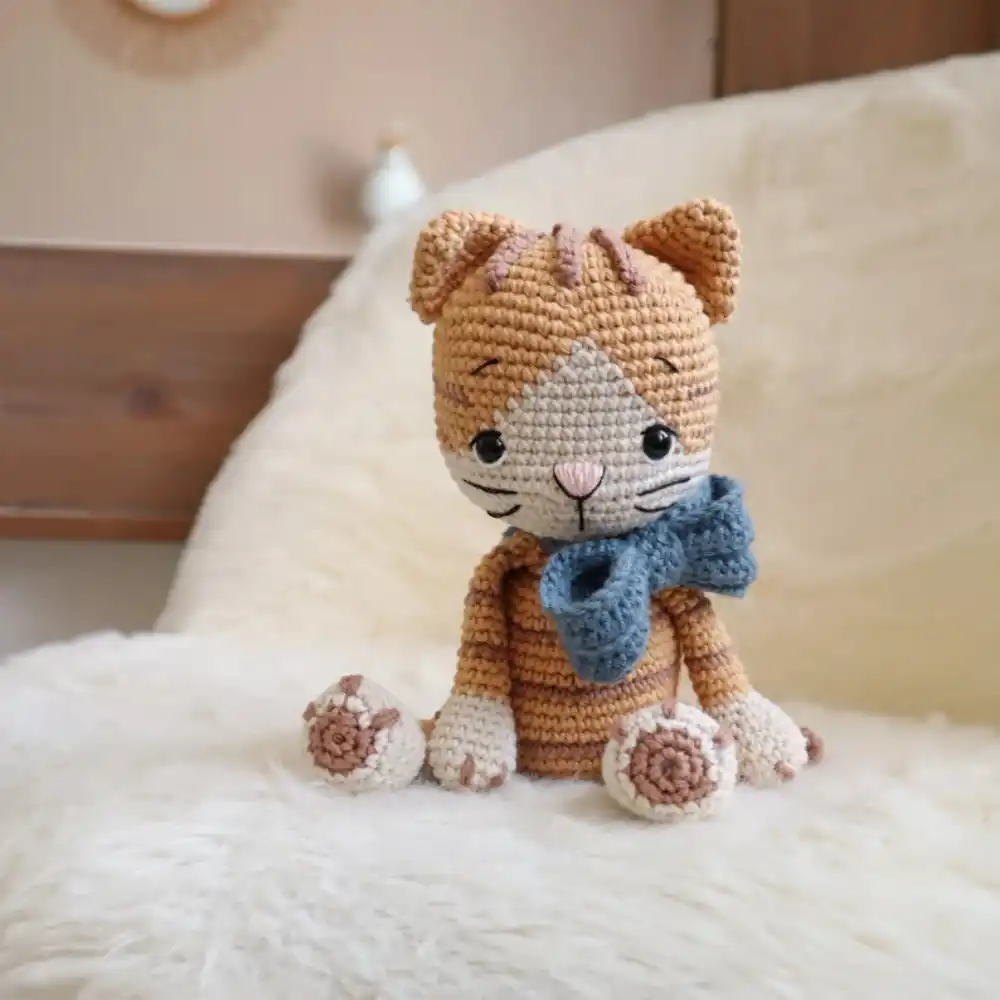 Gatinho tigrado em amigurumi caramelo com focinho branco, nariz rosa e laço azul