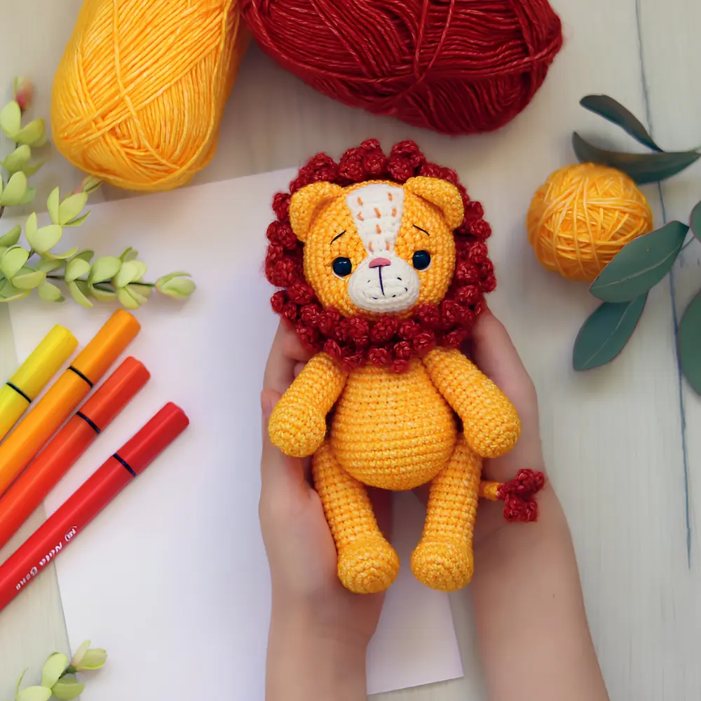 Leãozinho em amigurumi amarelo-alaranjado com juba vermelha, focinho claro e cauda