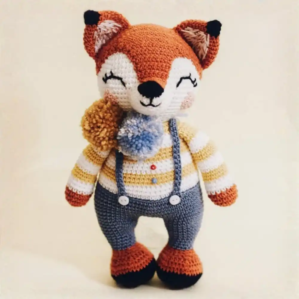 Raposinha em amigurumi laranja e branca com blusa listrada, calça cinza com suspensórios e pompons