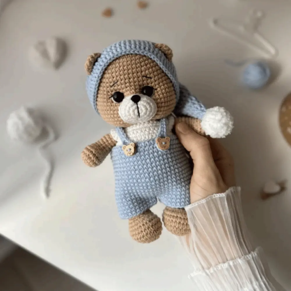 Ursinho marrom em amigurumi com macacão azul e touca comprida com pompom branco